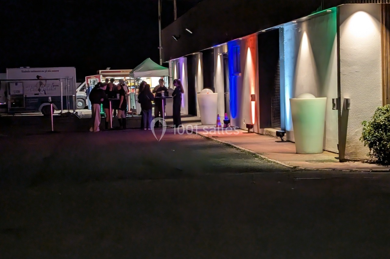 Groupe de personnes rassemblées près d'un stand éclairé, devant un bâtiment illuminé de lumières colorées la nuit.