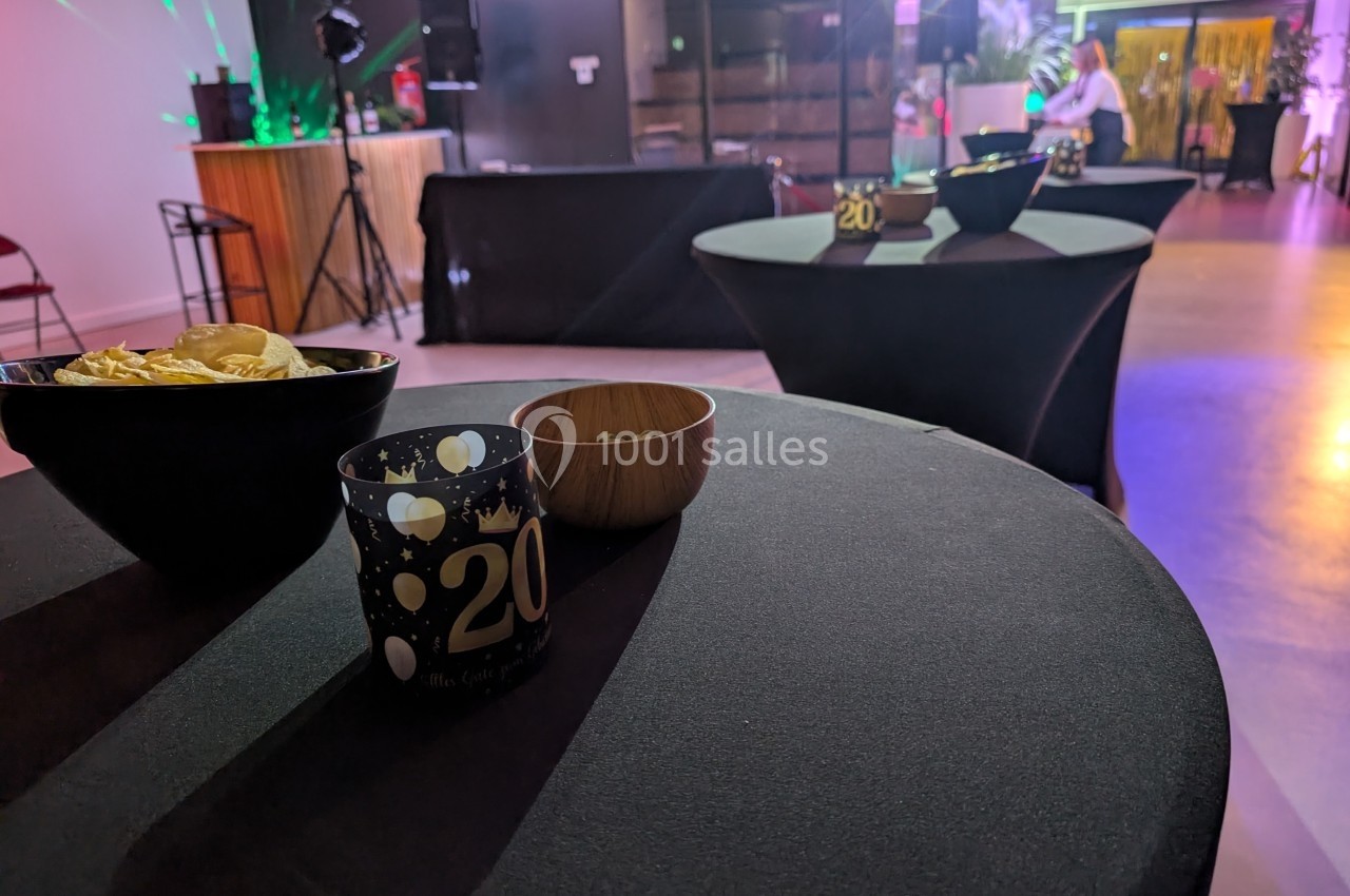 Tables hautes décorées pour une fête, avec bougies et bols, dans une salle éclairée par des lumières colorées.