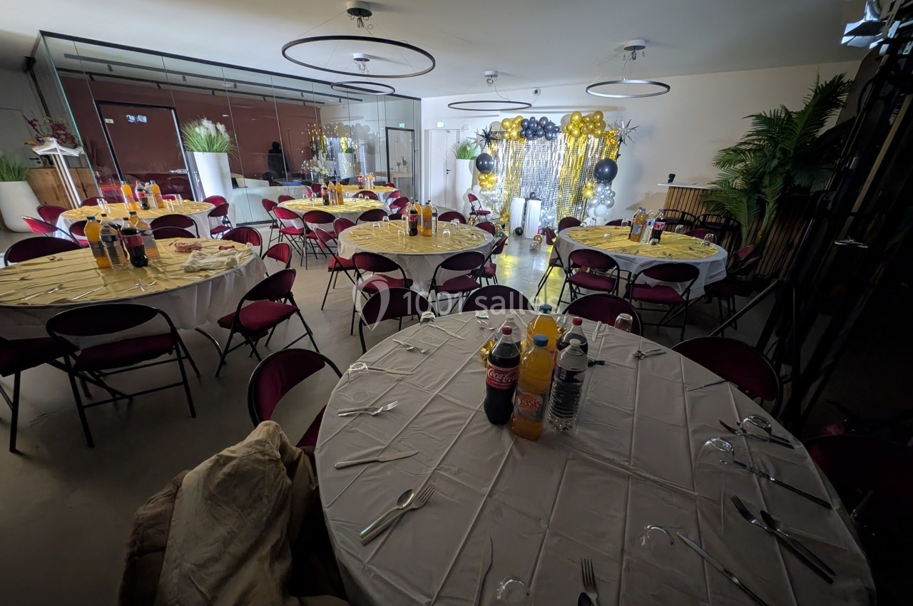 Salle décorée pour un événement avec tables rondes dressées, chaises rouges et arrière-plan orné de ballons et guirlandes.