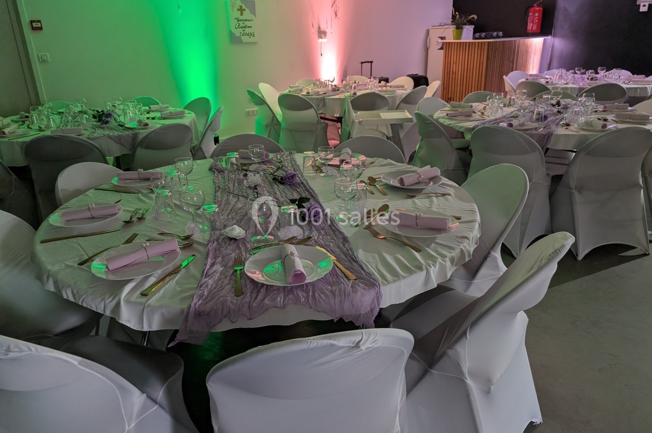 Salle de réception décorée avec des tables rondes, nappes blanches, chemins de table violets et vaisselle élégante.