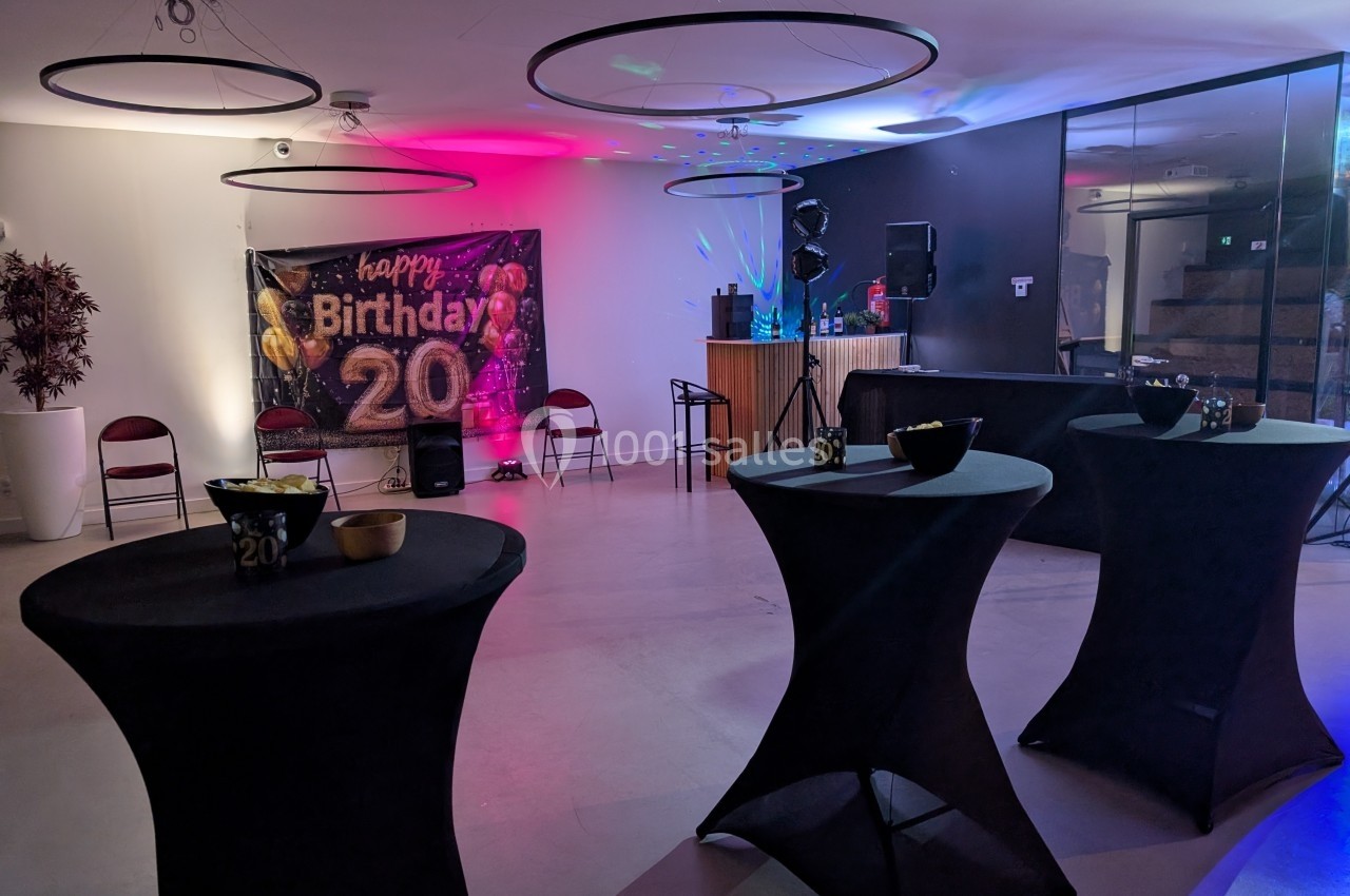 Salle décorée pour un 20e anniversaire avec tables hautes noires, éclairage coloré et une bannière festive.