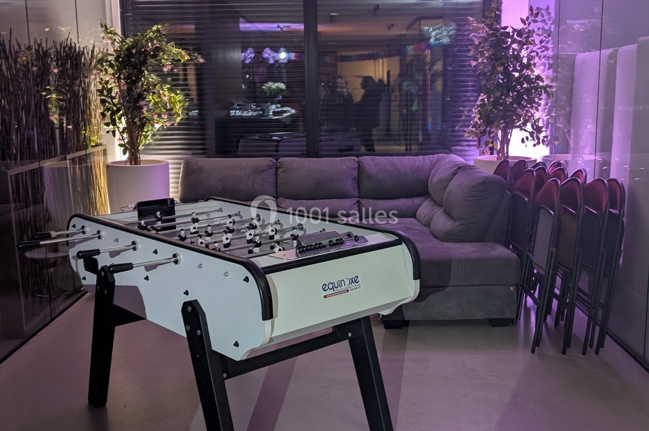 Table de baby-foot blanche devant un canapé gris, avec des chaises empilées et des plantes en arrière-plan.