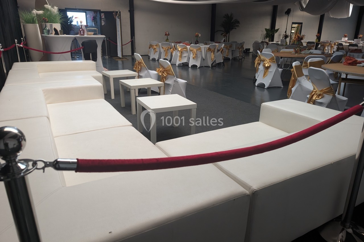 Salle de réception décorée avec des tables et chaises blanches ornées de nœuds dorés, et un espace lounge avec canapés…