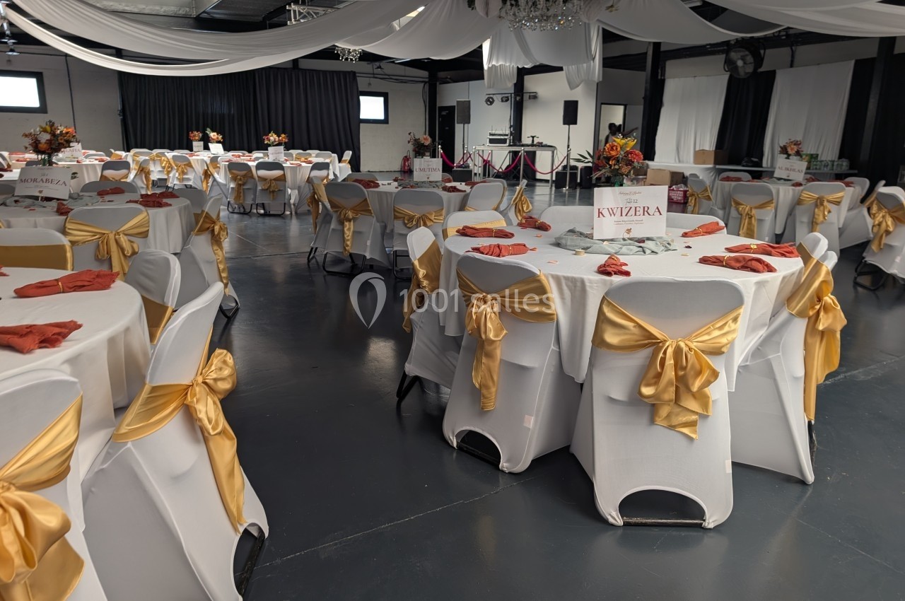 Salle de réception décorée avec des nappes blanches, des nœuds dorés sur les chaises et des tables ornées de fleurs.