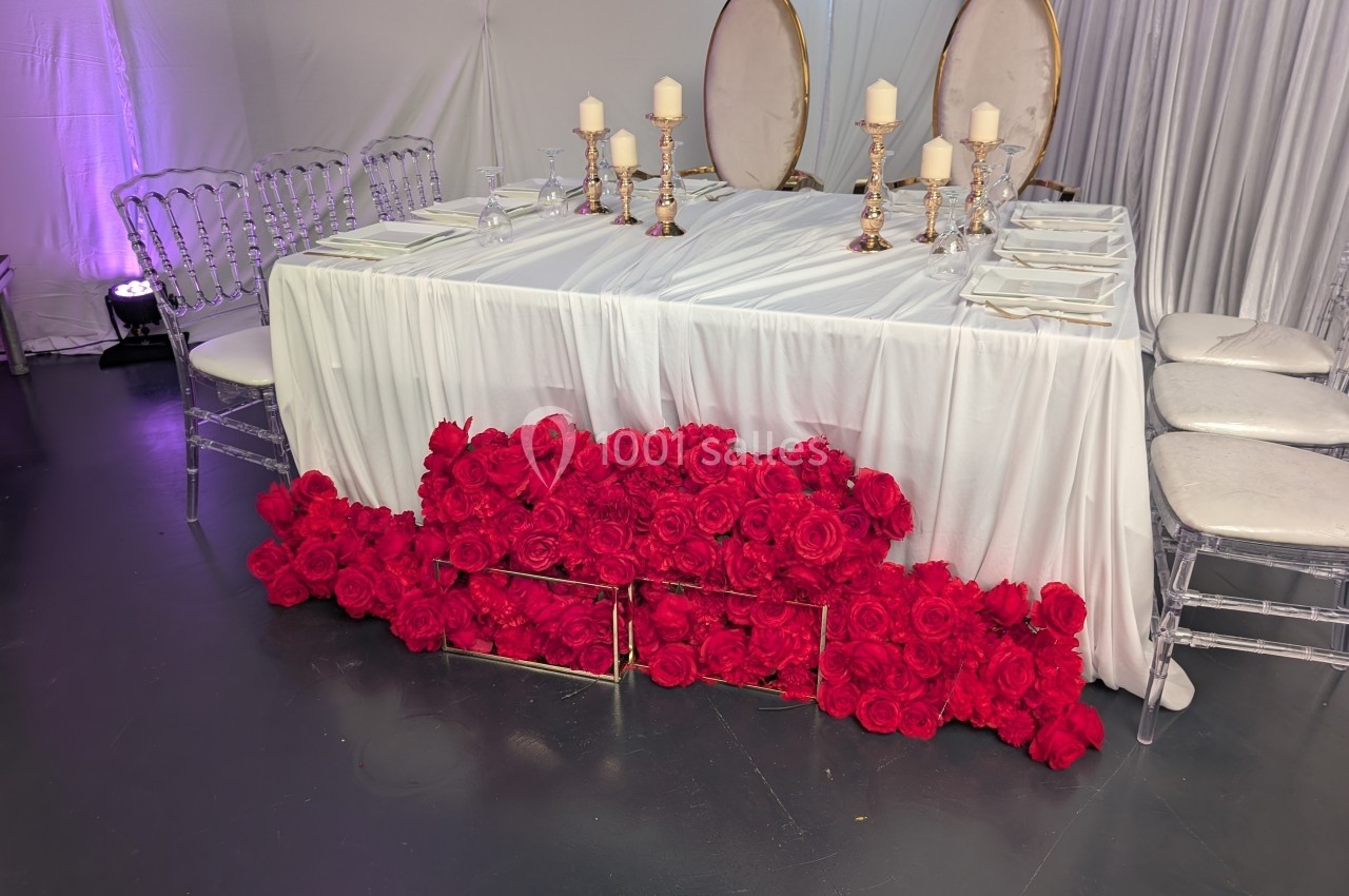 Table décorée avec une nappe blanche, chandeliers dorés, assiettes blanches et roses rouges disposées en bas.