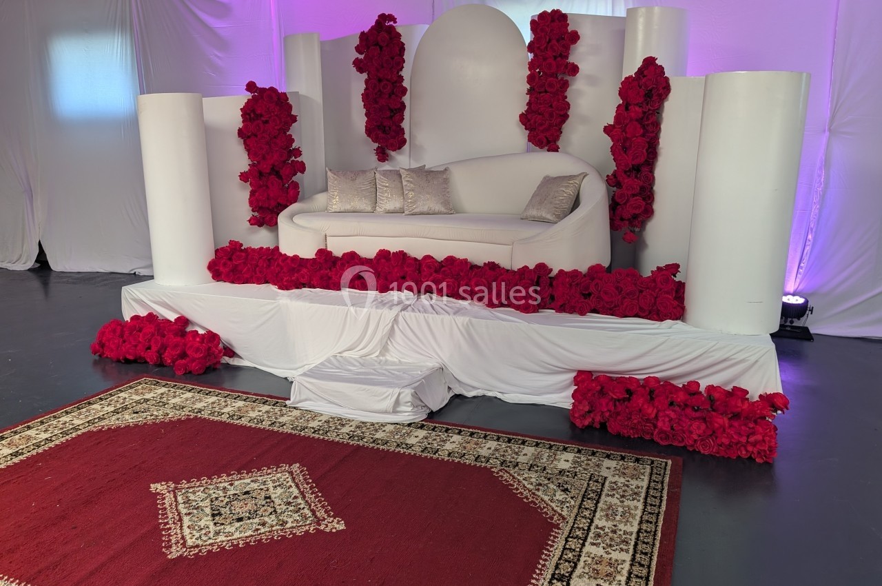 Canapé blanc décoré de roses rouges sur une estrade, avec un tapis rouge et blanc au sol dans un décor lumineux.