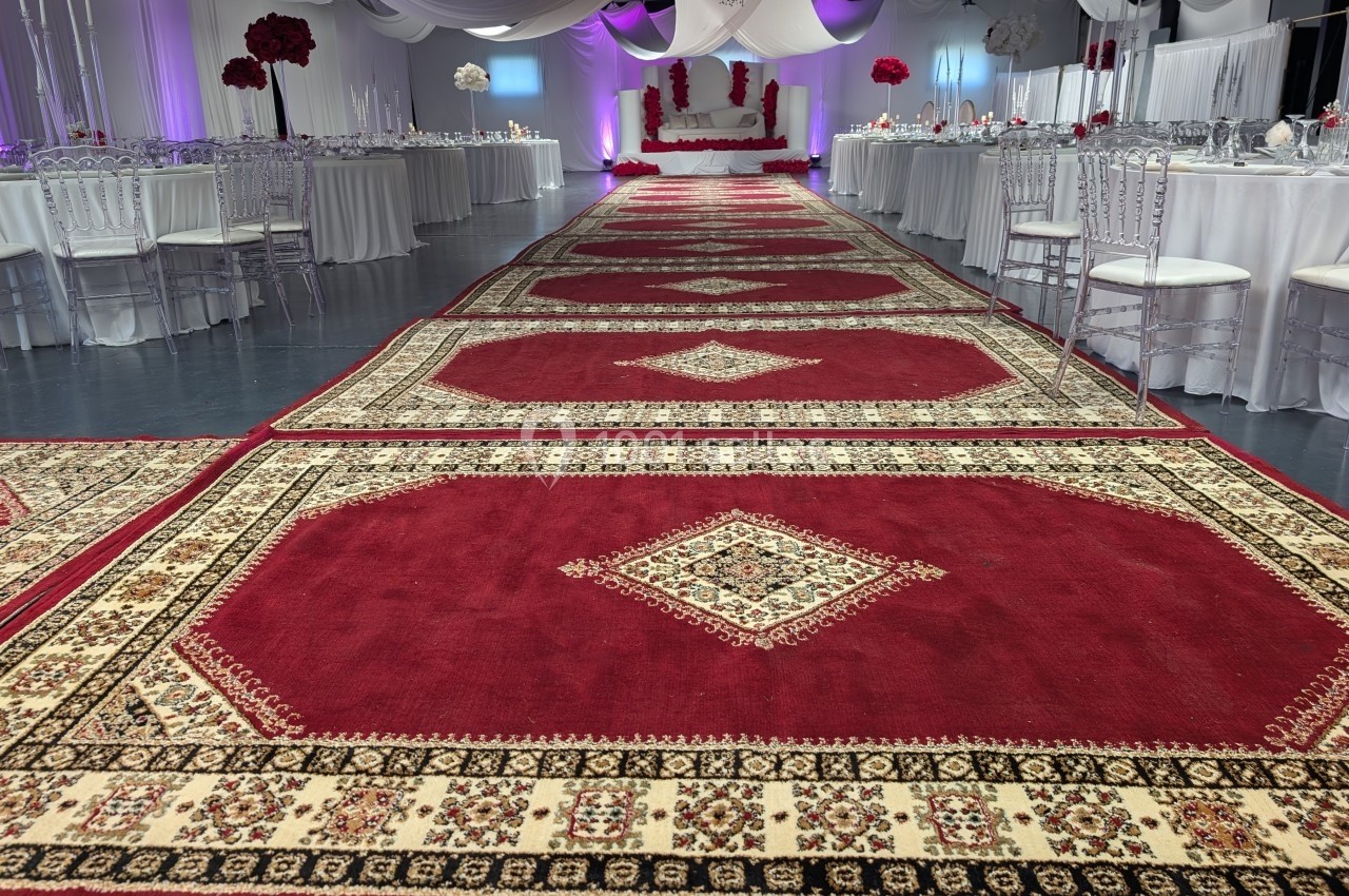 Salle de réception décorée avec des nappes blanches, tapis rouges ornés de motifs et une table d'honneur en arrière-plan.