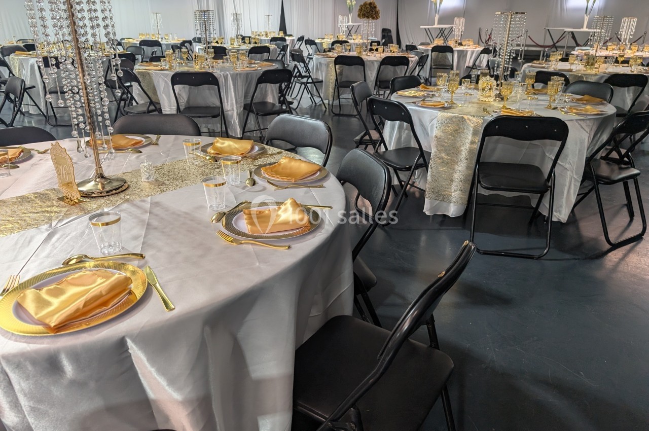 Salle de réception décorée avec des tables rondes dressées de nappes blanches, serviettes dorées et chaises noires.