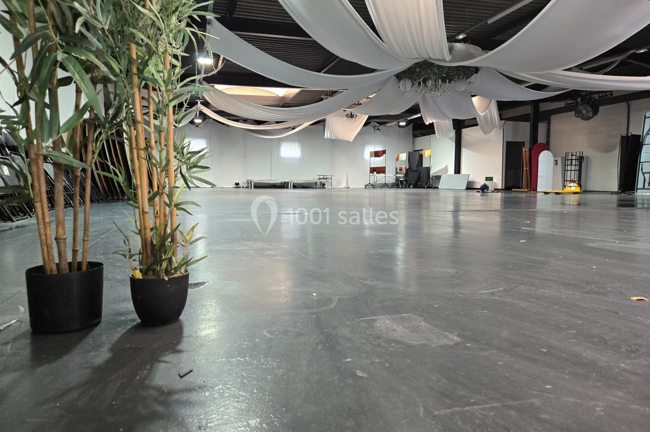 Grande salle vide avec sol en béton, décorée de drapés blancs suspendus au plafond et plantes en pot au premier plan.