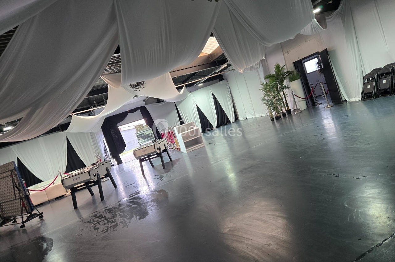 Grande salle avec sol sombre, plafond décoré de drapés blancs, baby-foot et plantes en arrière-plan.
