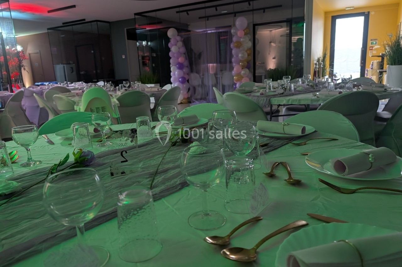 Salle de réception décorée avec des tables dressées, nappes blanches, couverts dorés et ballons en arrière-plan.