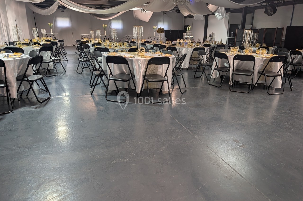 Salle de réception décorée avec des tables rondes dressées, nappes blanches et chaises noires, sous des drapés suspendus.