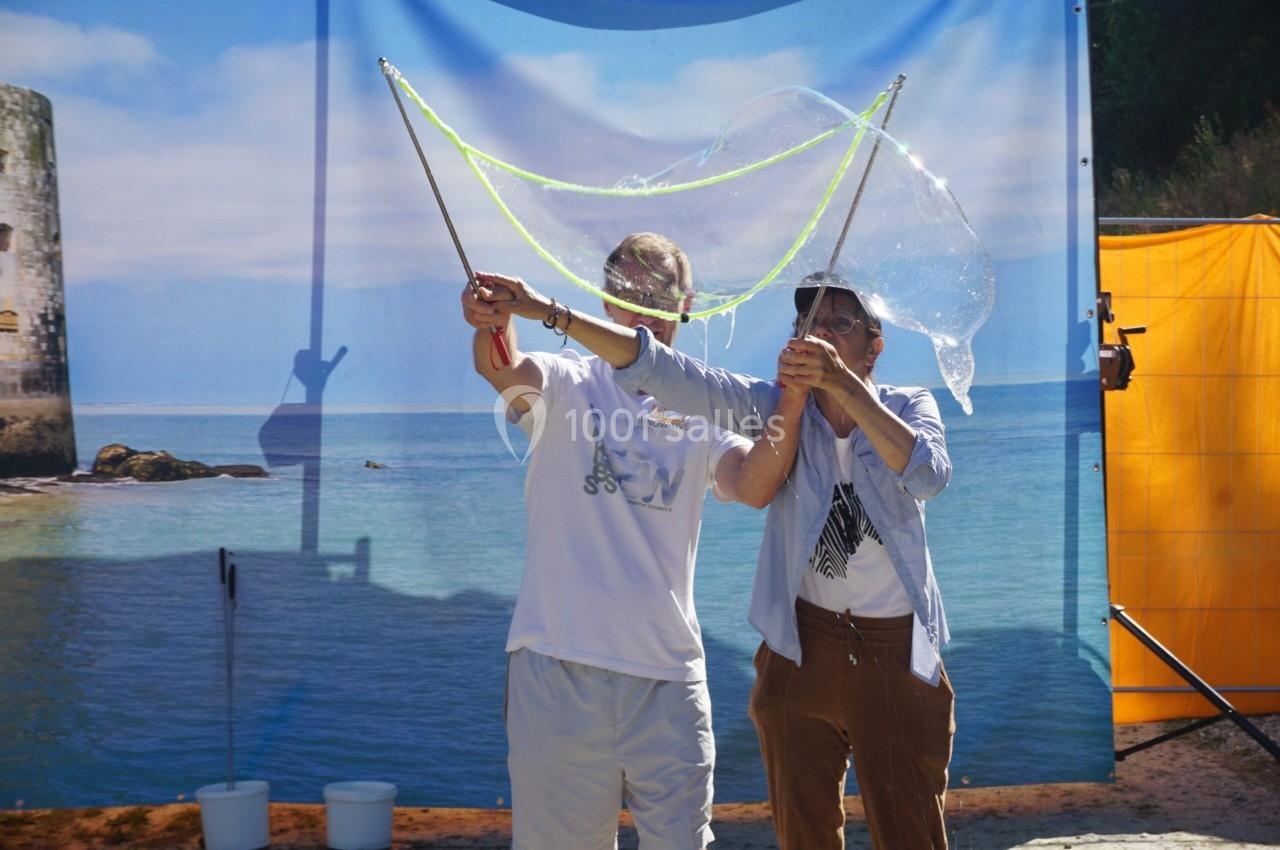 Deux personnes créent une grande bulle de savon à l'aide de baguettes devant un décor de plage.