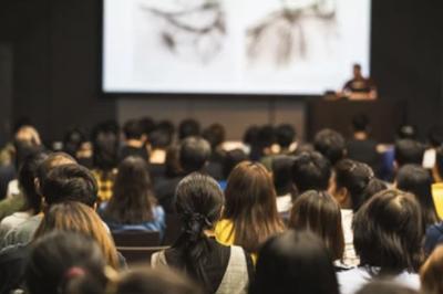 Un grand groupe de jeunes assis dehors sur un sol en béton, près de bâtiments sous un ciel nuageux.