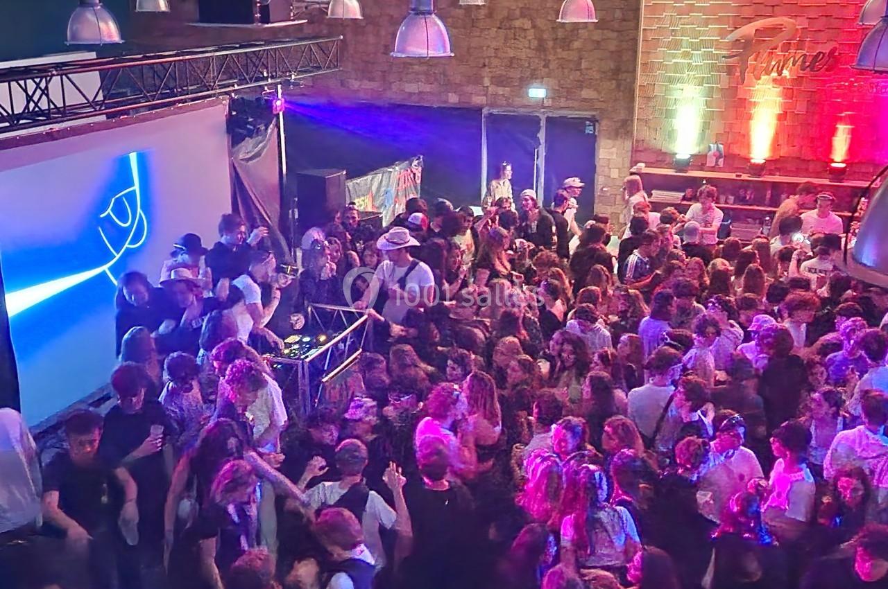 Foule dansante dans une salle éclairée par des lumières colorées, avec un DJ sur scène et un écran lumineux en arrière-plan.