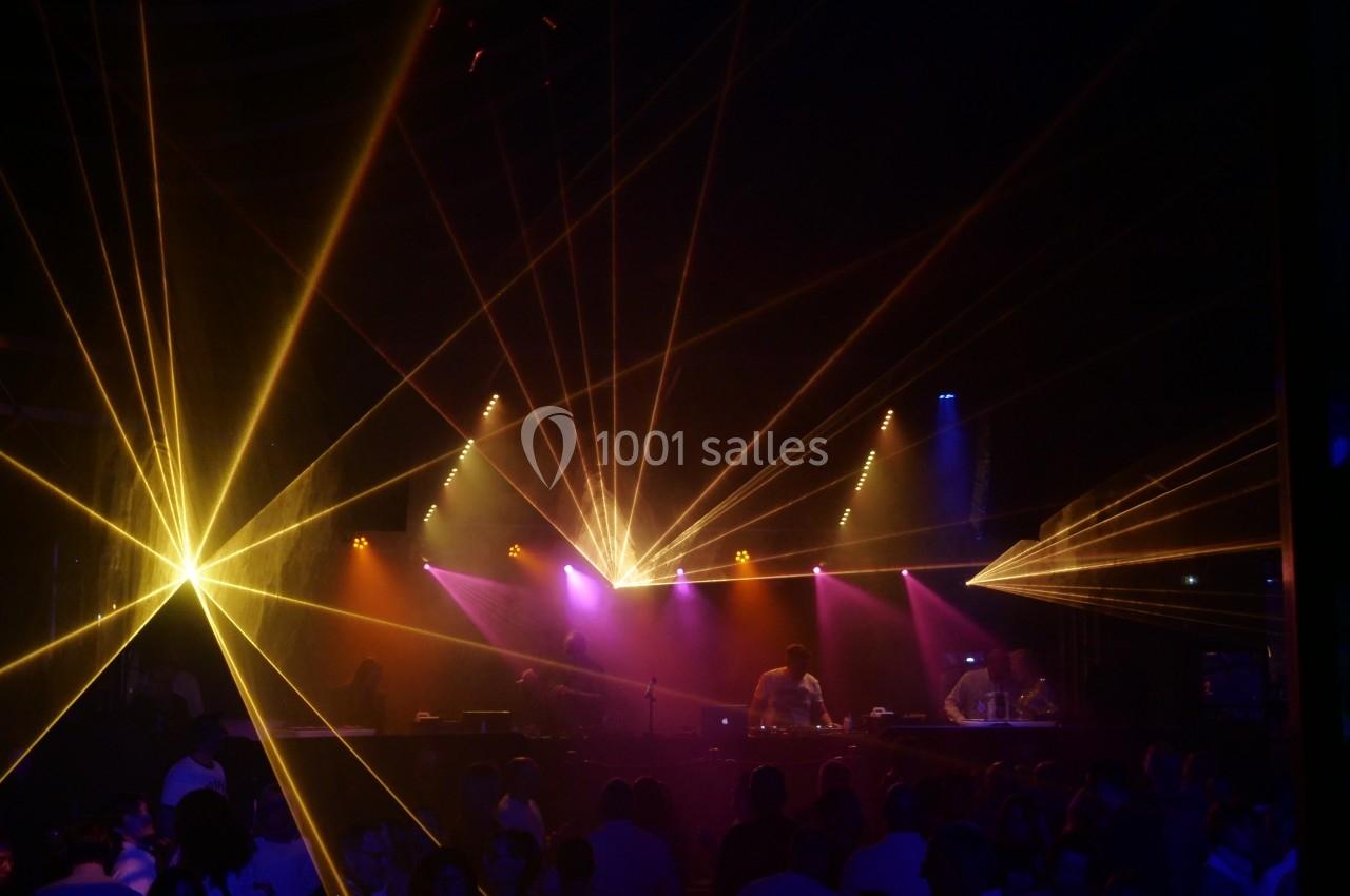 Salle sombre avec des jeux de lumières colorées et des faisceaux laser, des personnes dansent devant une scène.