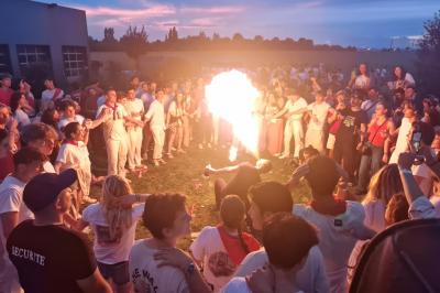 Un grand groupe de jeunes assis dehors sur un sol en béton, près de bâtiments sous un ciel nuageux.