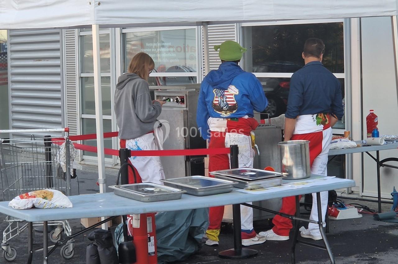 Trois personnes en tabliers préparent de la nourriture sur un stand extérieur équipé de plaques de cuisson et de tables.