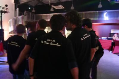 Un grand groupe de jeunes assis dehors sur un sol en béton, près de bâtiments sous un ciel nuageux.