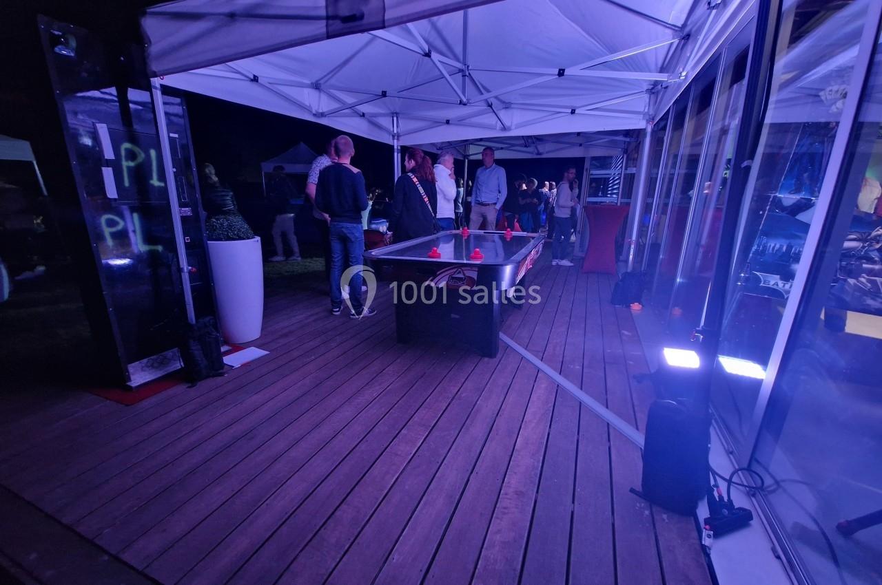 Des personnes discutent sous des tentes éclairées par des lumières violettes, sur une terrasse en bois avec une table de jeu.