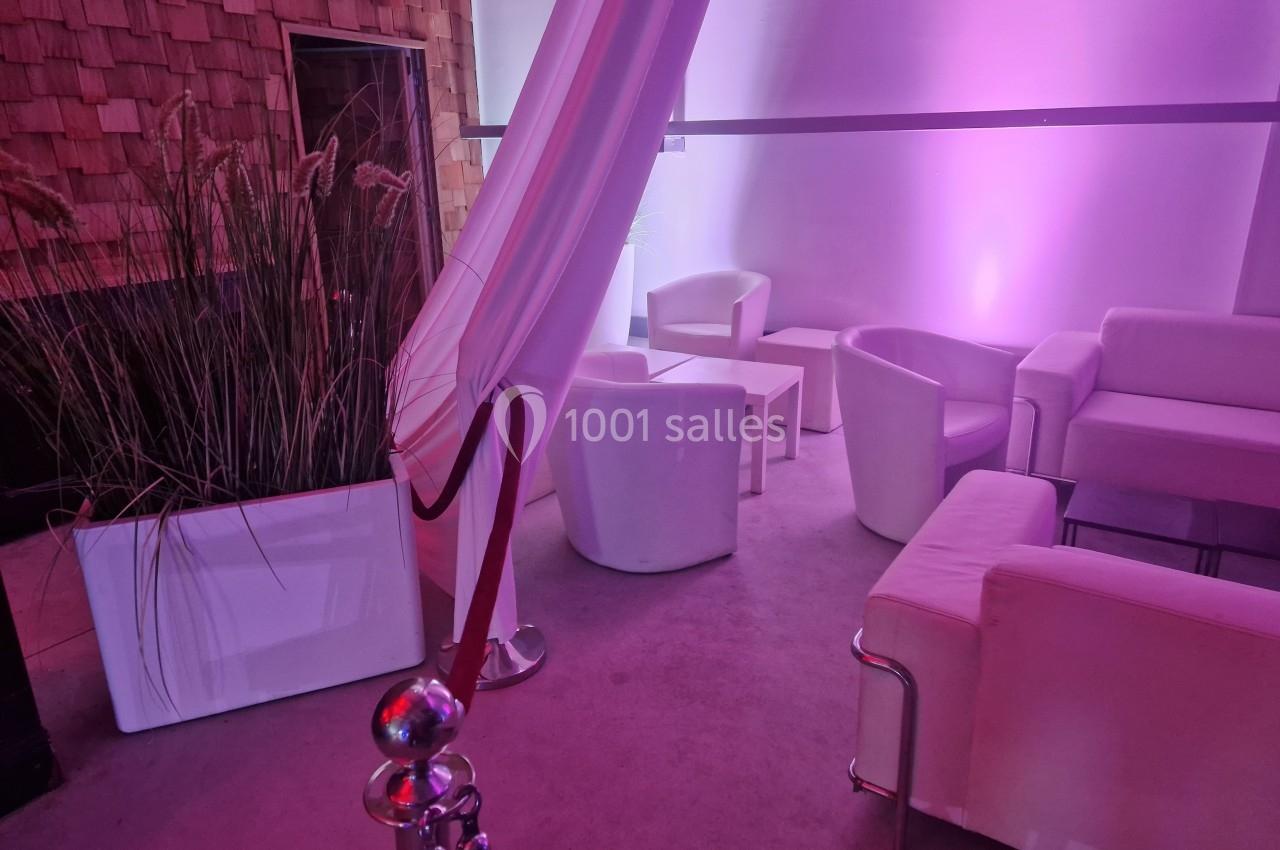 Espace lounge avec fauteuils et canapés blancs, éclairé par une lumière rose, décoré de plantes en pot.