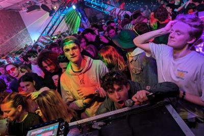 Un grand groupe de jeunes assis dehors sur un sol en béton, près de bâtiments sous un ciel nuageux.