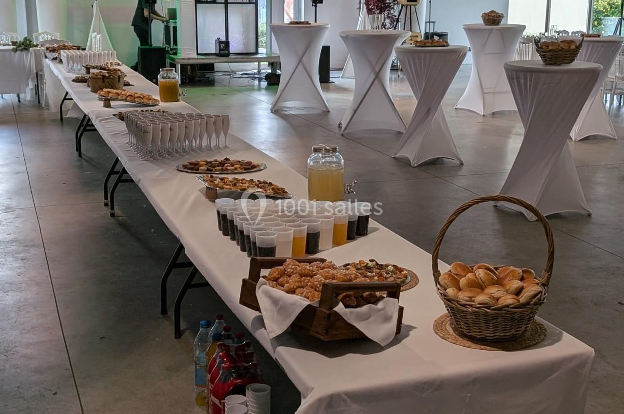 Table dressée avec viennoiseries, pains, jus et verres, dans une salle lumineuse avec tables hautes en arrière-plan.