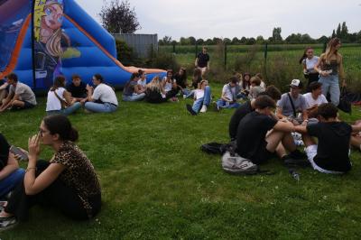 Un grand groupe de jeunes assis dehors sur un sol en béton, près de bâtiments sous un ciel nuageux.