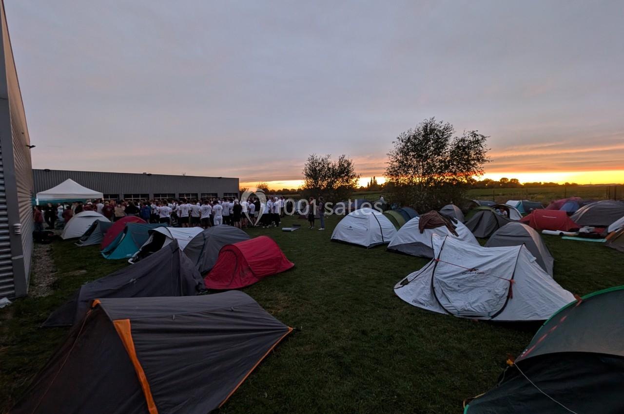 Tentes installées sur une pelouse au coucher du soleil, avec un groupe de personnes rassemblées près d'un bâtiment.