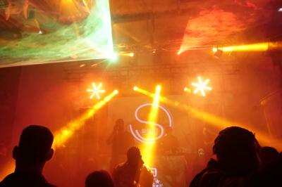 Un grand groupe de jeunes assis dehors sur un sol en béton, près de bâtiments sous un ciel nuageux.