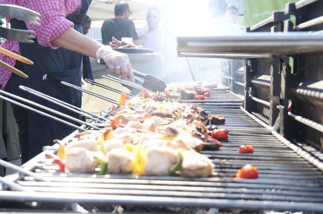 Personnes cuisinant des brochettes de viande et légumes sur un barbecue en plein air, avec de la fumée visible.