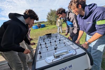 Un grand groupe de jeunes assis dehors sur un sol en béton, près de bâtiments sous un ciel nuageux.