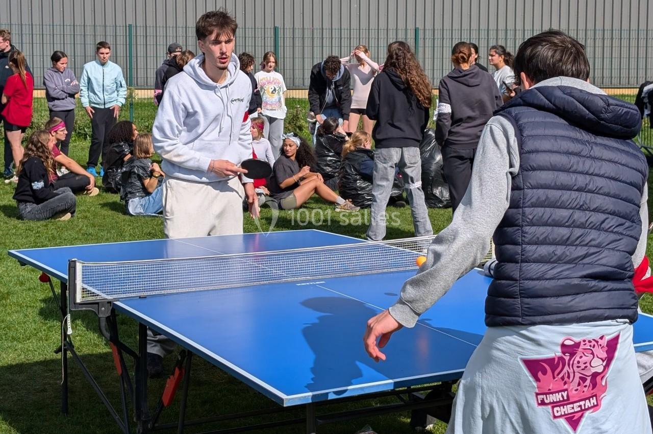 Deux personnes jouent au tennis de table en extérieur, entourées de spectateurs assis ou debout sur une pelouse.
