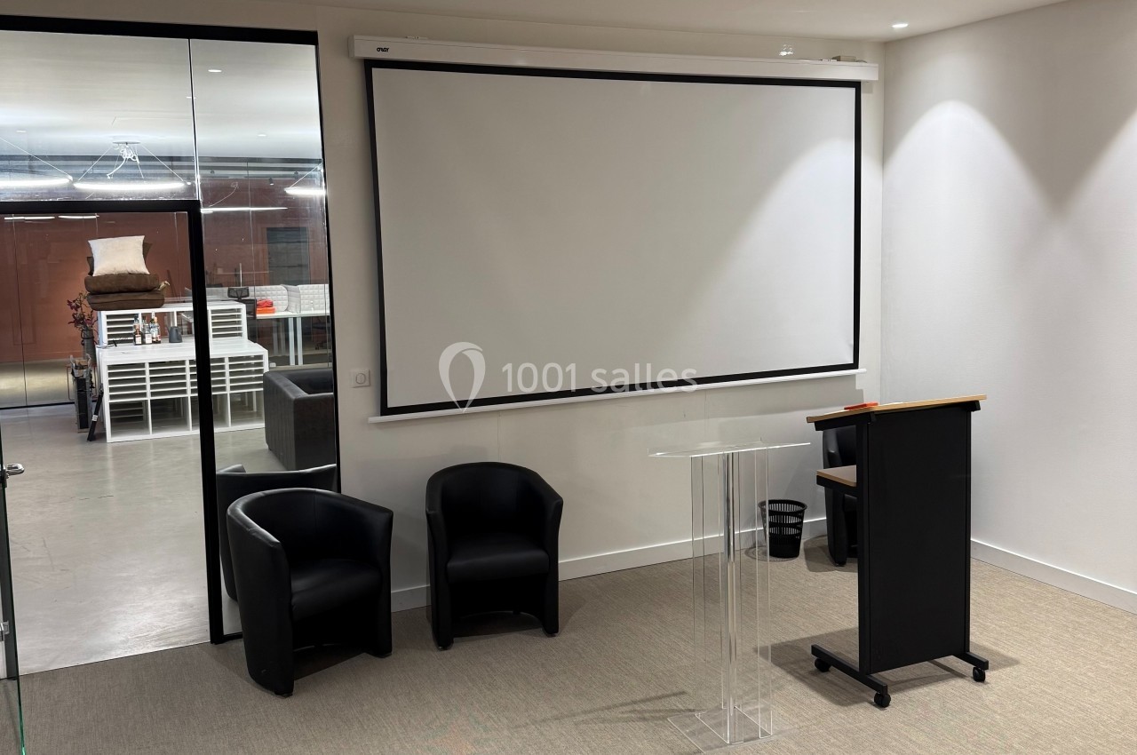 Salle de réunion avec écran de projection, pupitre transparent, deux fauteuils noirs et vue sur un espace adjacent.