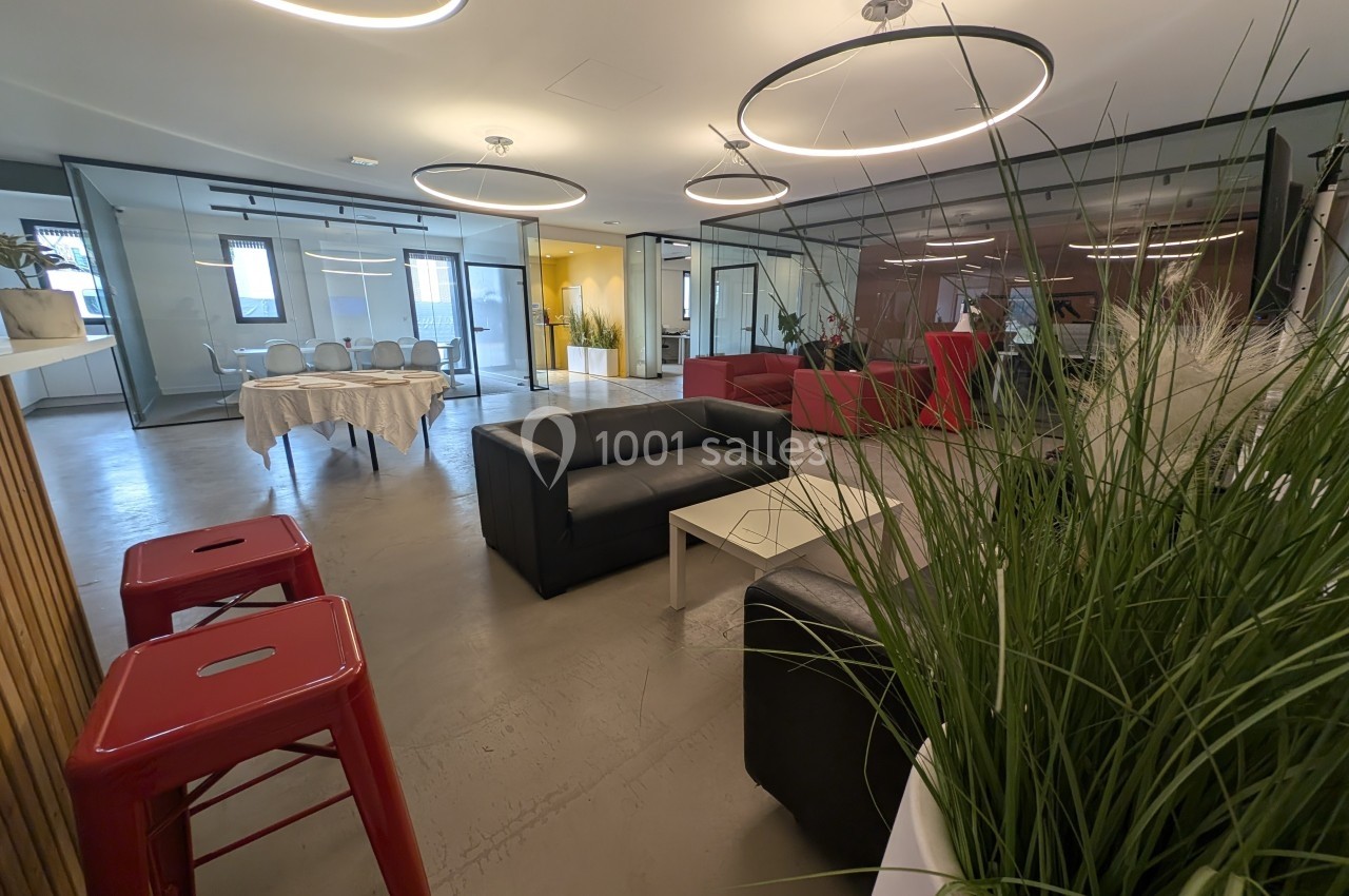 Espace intérieur moderne avec canapés noirs, tabourets rouges, plantes et grandes baies vitrées.