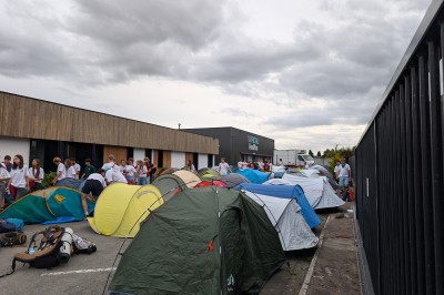 Un grand groupe de jeunes assis dehors sur un sol en béton, près de bâtiments sous un ciel nuageux.