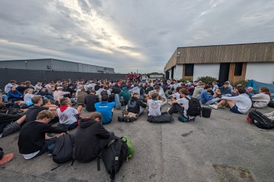 Un grand groupe de jeunes assis dehors sur un sol en béton, près de bâtiments sous un ciel nuageux.