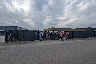 Un grand groupe de jeunes assis dehors sur un sol en béton, près de bâtiments sous un ciel nuageux.