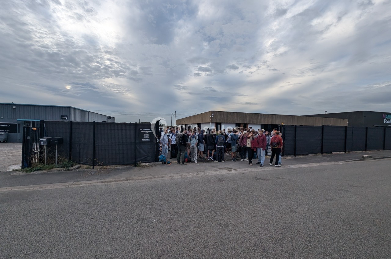 Un groupe de personnes rassemblées devant une clôture noire dans une zone industrielle sous un ciel nuageux.