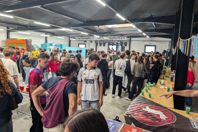 Un grand groupe de jeunes assis dehors sur un sol en béton, près de bâtiments sous un ciel nuageux.