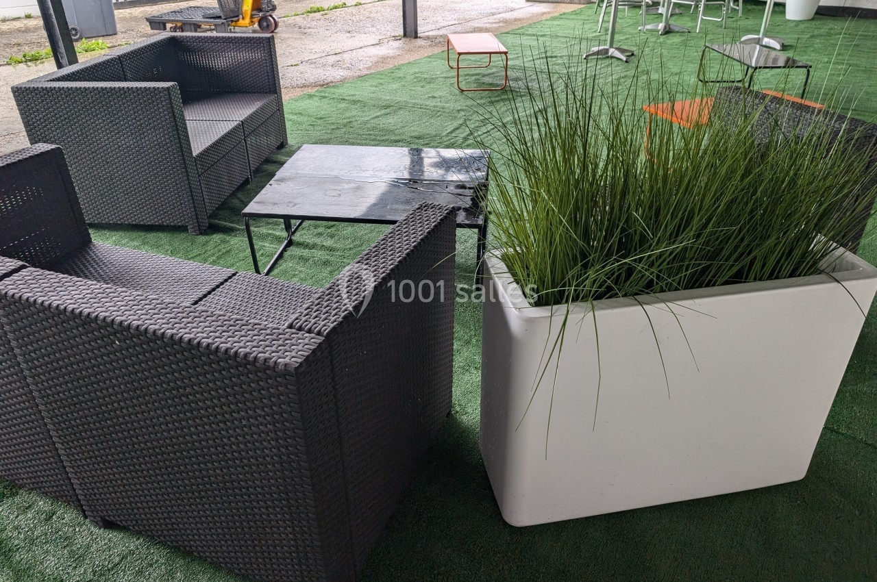 Mobilier d'extérieur avec fauteuils en résine tressée, tables basses noires et jardinière blanche sur gazon synthétique.