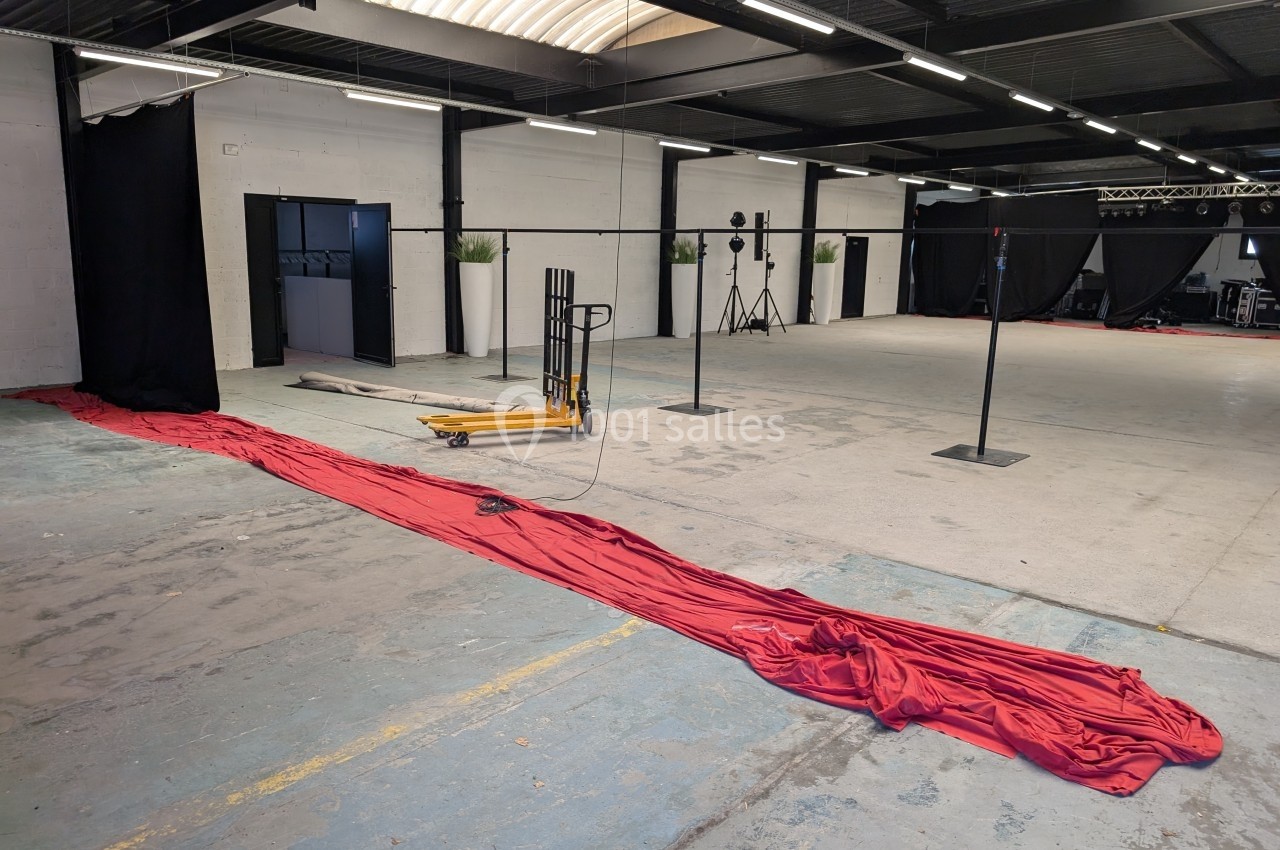 Grand espace intérieur industriel avec sol en béton, rideau rouge étendu au sol et équipements de studio en arrière-plan.