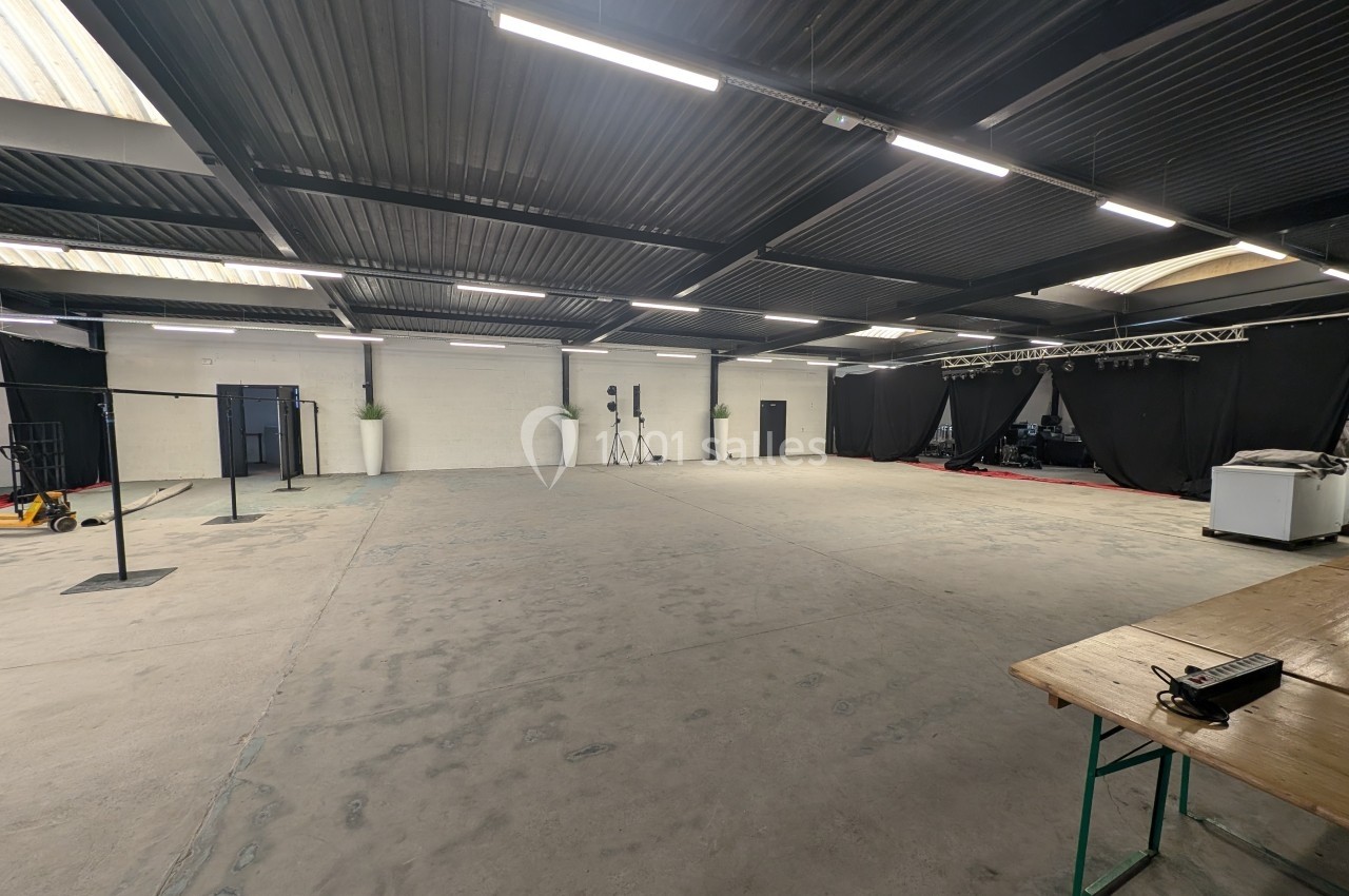 Grande salle vide avec sol en béton, plafond métallique, éclairage suspendu et scène équipée de rideaux noirs au fond.