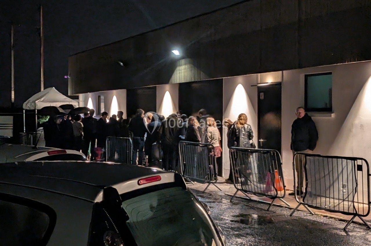 Des personnes font la queue sous la pluie devant un bâtiment éclairé, délimité par des barrières métalliques.