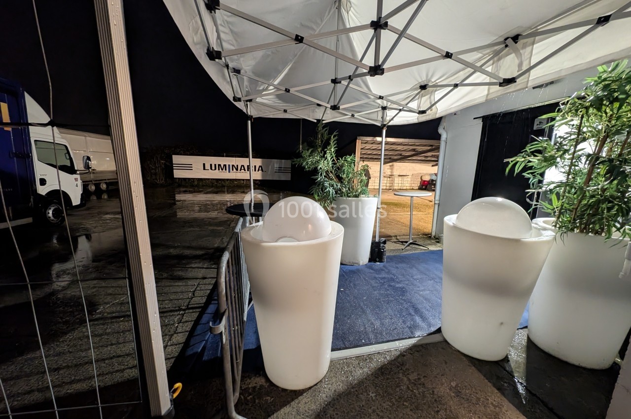 Vue nocturne d'un espace extérieur couvert par une tente, avec des plantes et des luminaires décoratifs.