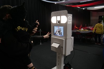 Des enfants colorient des dessins autour d'une table ronde, entourés d'adultes dans un espace intérieur animé.