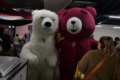 Des enfants colorient des dessins autour d'une table ronde, entourés d'adultes dans un espace intérieur animé.