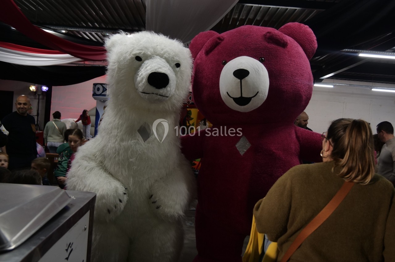 Deux mascottes en ours, blanc et rose, posent debout dans un espace intérieur animé avec des visiteurs.