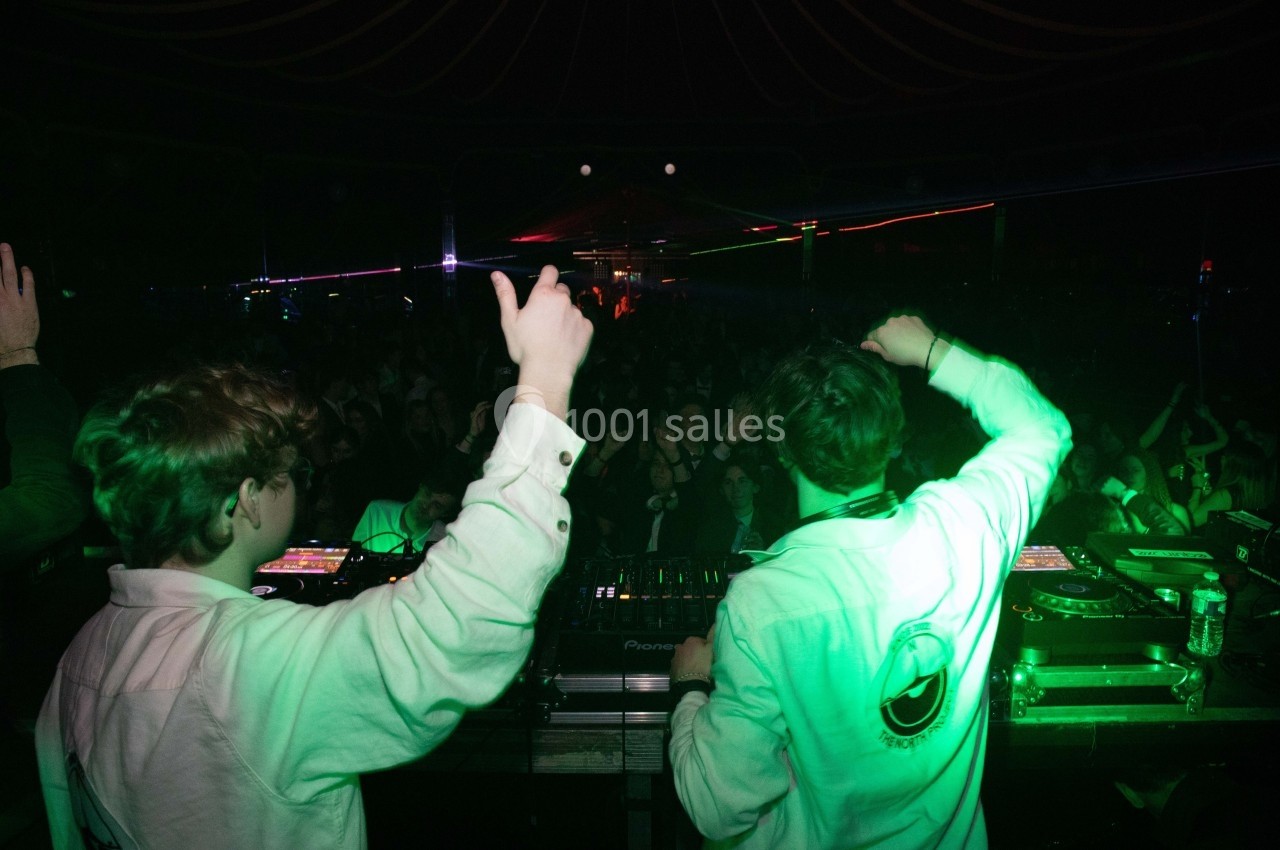 Deux DJ en chemises blanches mixent sur scène devant une foule dans une ambiance lumineuse de concert.