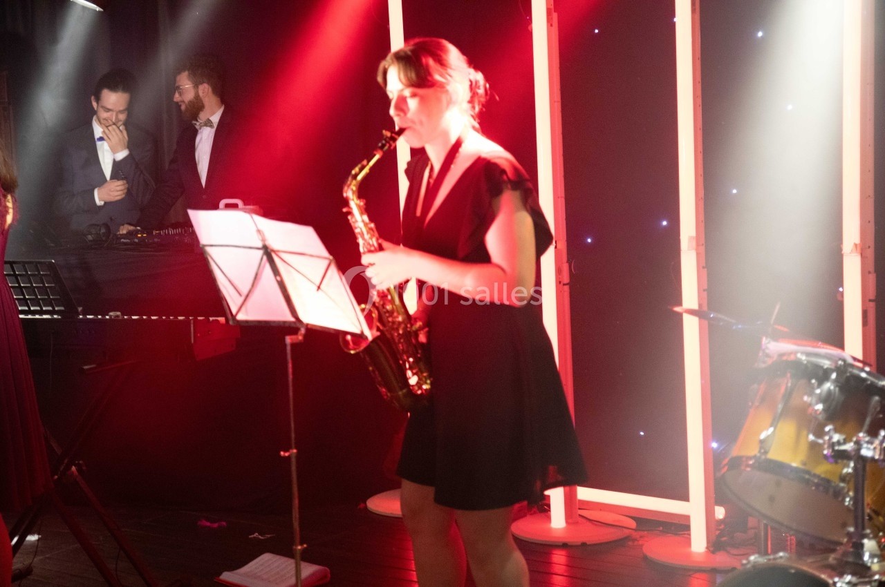 Une femme joue du saxophone sur scène, éclairée par des lumières rouges, avec des musiciens en arrière-plan.